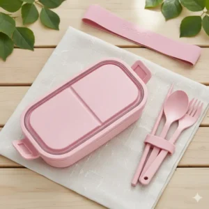 Pink bento lunch box displayed next to its reusable bamboo cutlery set and matching carrying strap.